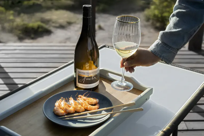 Hand holding glass of white wine from Amelia bottle beside grilled shrimp on plate, on outdoor tray