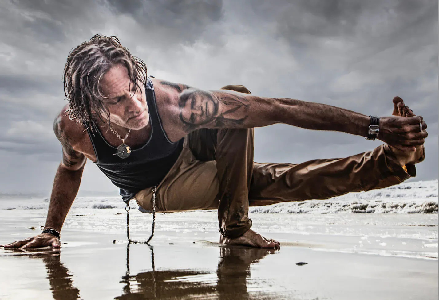 Eric Paskel performs yoga pose on beach, muscular tattooed man in tank top and pants, dramatic cloudy sky, wet sand reflection.
