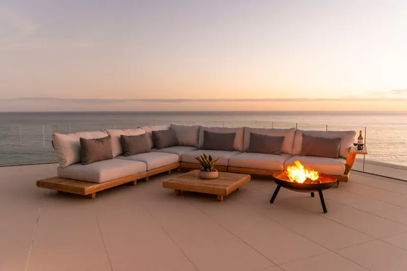 Beige L-shaped wooden outdoor sofa and fire pit on terrace overlooking ocean at sunset