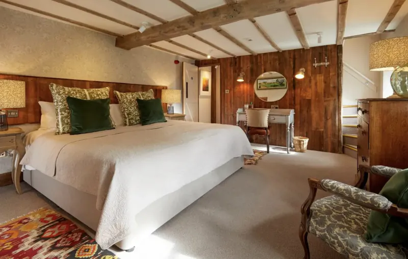 Cozy Cotswolds bedroom at The Crown Inn with wooden beams, king bed with green pillows, desk, armchair, and rug.