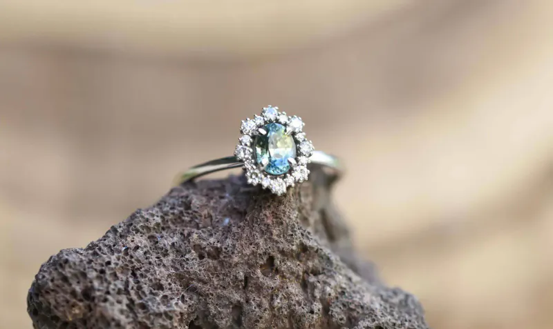 Oval teal gemstone ring with diamond halo on rugged rock surface