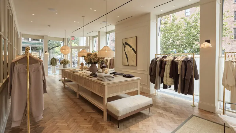 Spacious Varley flagship store interior in SoHo with wooden table display, racks of neutral clothing, pendant lights, and large windows.