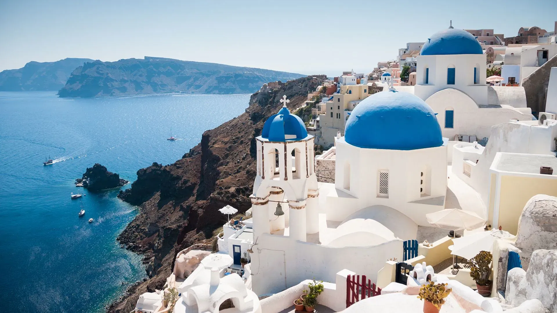 Iconic white Oia buildings with blue domes perched on Santorini cliff overlooking turquoise Aegean Sea and sailboats.