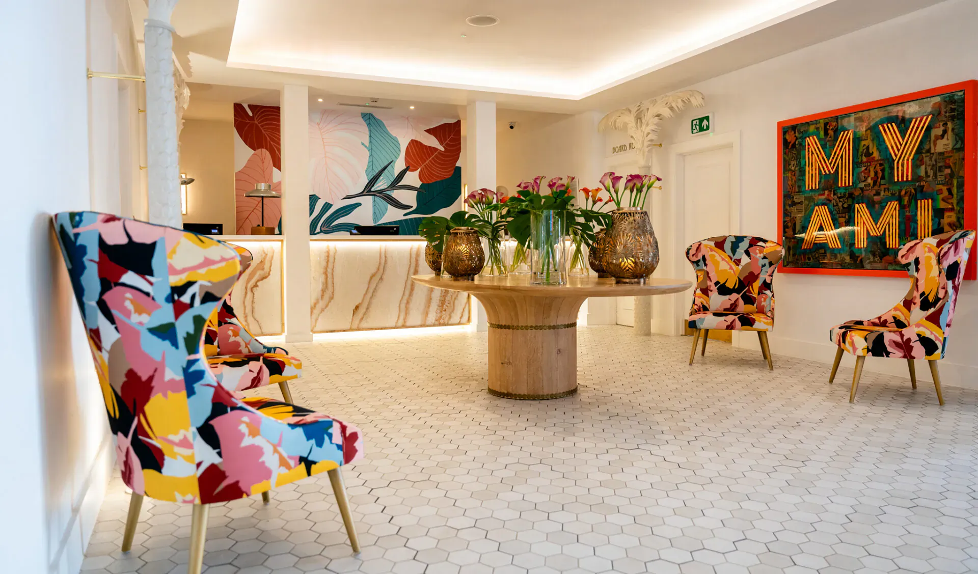 Stylish hotel lobby with colorful patterned armchairs around wooden table, plants, 'MY AMI' artwork, marble reception.