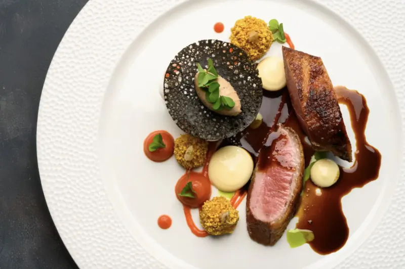 Elegant plate of seared duck breast with black tuile, quail egg, herb crisp, tomato dots, and jus at De Tafel Restaurant