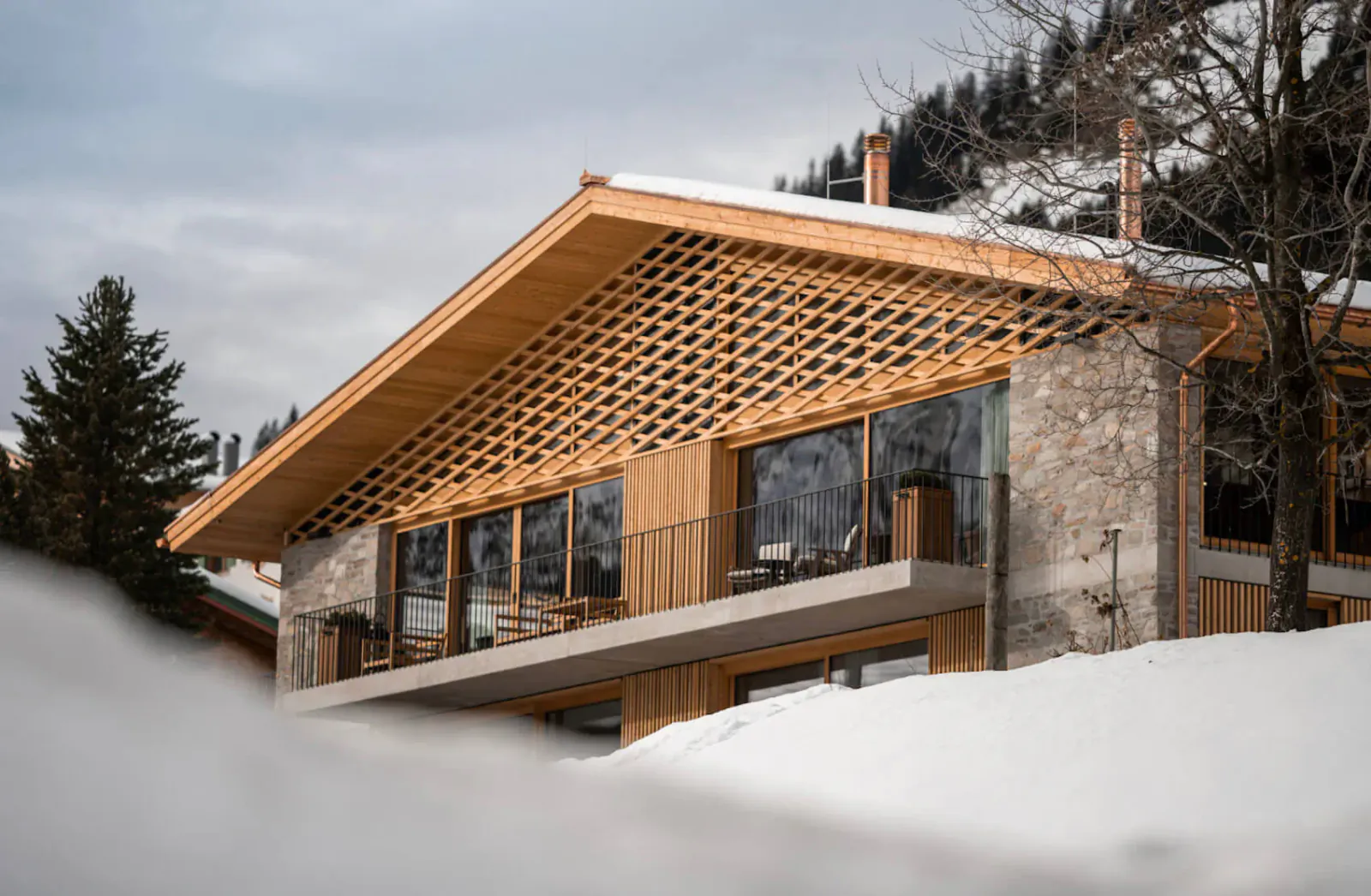 Stylish Austrian chalet with wooden lattice facade, large windows, balcony, and snow-covered alpine surroundings.