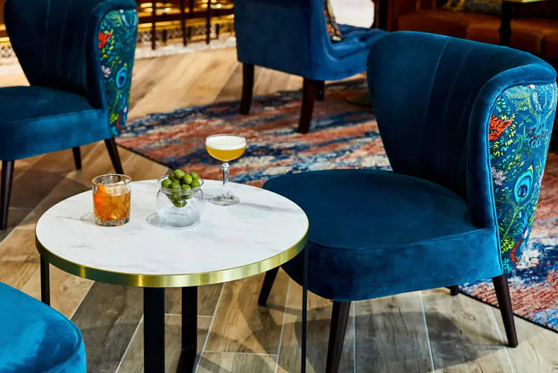 Cozy seating area in Hotel Indigo Chester with blue patterned velvet chairs around a marble table holding orange cocktail, grape spritz, and plant.