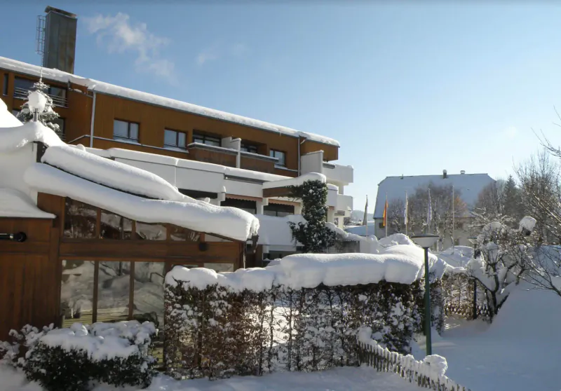 Snow-covered wooden building and garden at Karma Bavaria, Schliersee, in winter wonderland, clear blue sky.
