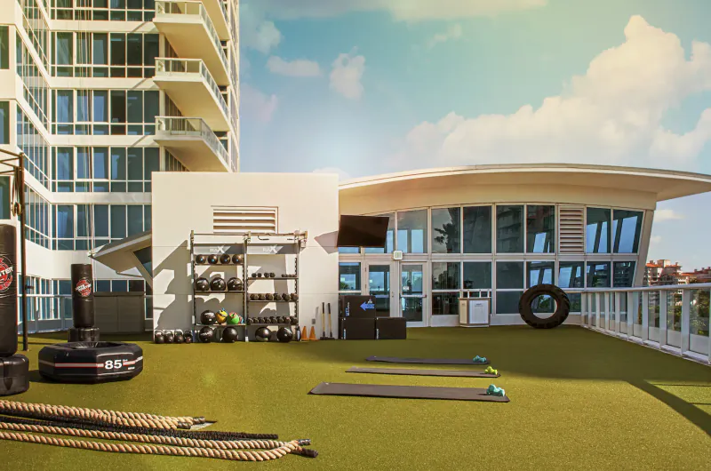 Modern rooftop gym at Continuum with turf, weights, mats, punching bags, tire, ropes, and tall glass condo building backdrop.