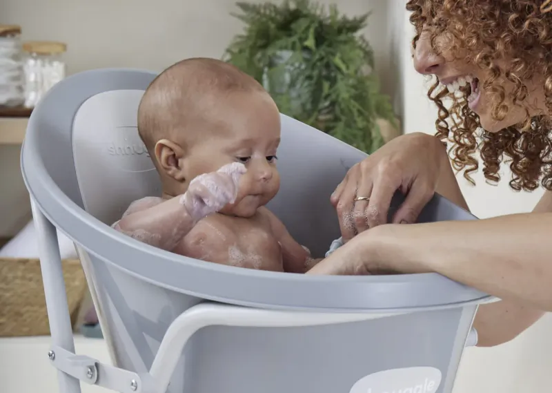 Smiling curly-haired mother bathing her baby in a gray tub, surrounded by plants.