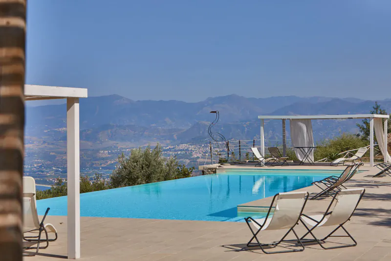 Infinity pool with loungers at luxury villa in Sicily, overlooking mountains and city under blue sky