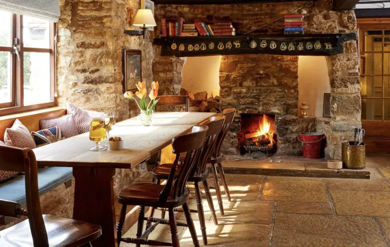 Cozy stone-walled inn dining area with wooden table, yellow tulips, cushioned bench, chairs by blazing fireplace.