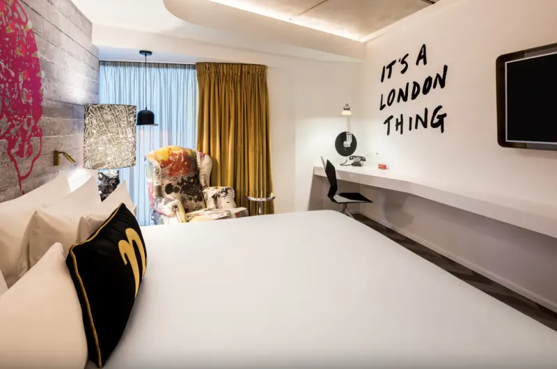 nhow London hotel room with 'It's a London Thing' wall graffiti, patterned armchair, desk, and bed with black pillows.