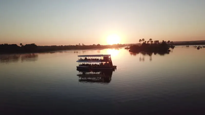 Double-decker boat on Zambezi River at sunset near Victoria Falls, with palm-covered islands and reflections.