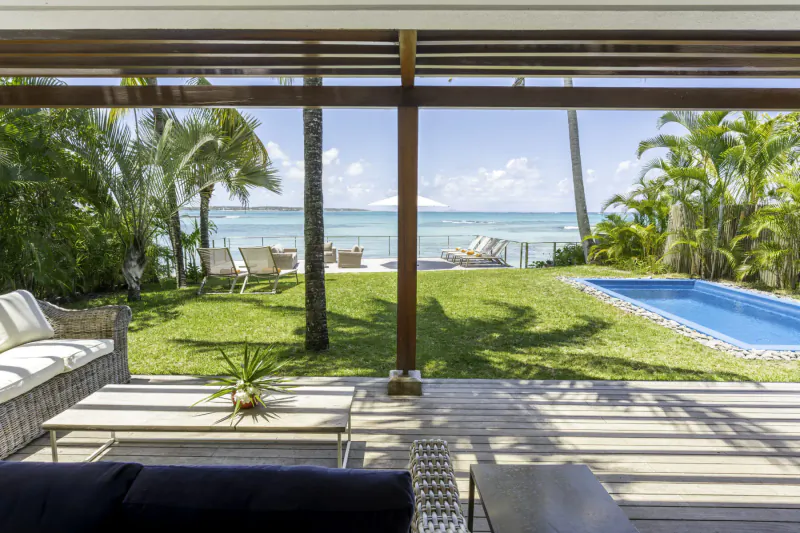Open wooden terrace of Villa Ruby, Mauritius, with white sofas, pool, lawn, palms, and Belle Mare beach view.