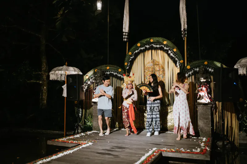 Balinese cultural performers in traditional attire on a flower-edged wooden stage at Kepitu Restaurant, night event.