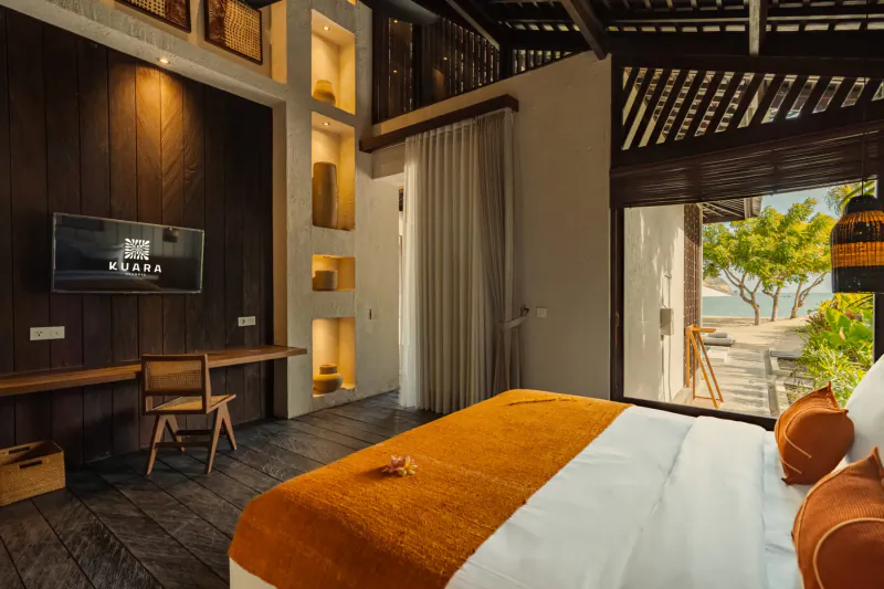 Luxurious bedroom in Kuara Lombok resort with orange bed, wooden decor, and beach view through open doors.