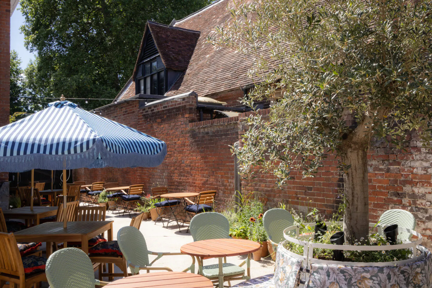 Outdoor seating area at The George & Dragon Marlow with blue umbrella, wooden tables, chairs, olive tree, brick walls, and riverside garden charm.