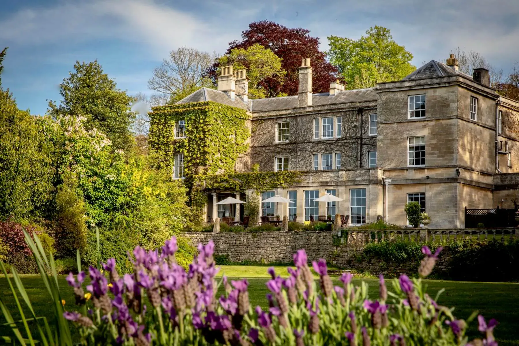 Burleigh Court, a ivy-covered stone manor in the Cotswolds, surrounded by trees and purple flowers in the foreground.