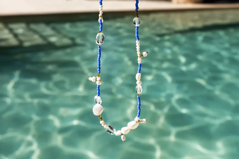 Blue beaded necklace with white pearls and clear pebble beads, hanging over turquoise pool water