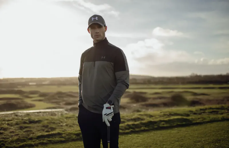 Man in Under Armour cap and gray zip-up jacket holds golf glove on course at sunset