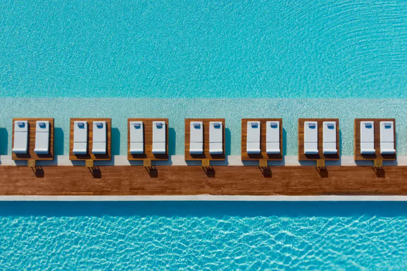 Aerial view of white cushioned lounge chairs lined up on a wooden deck beside a turquoise pool.