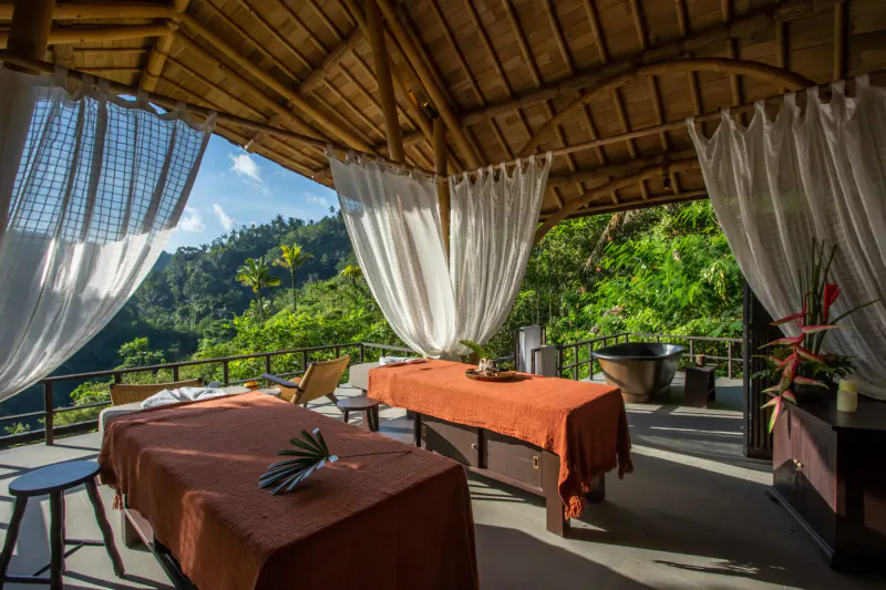 Open-air bamboo spa pavilion with two massage tables, white curtains, tropical plants, and lush Bali jungle view.