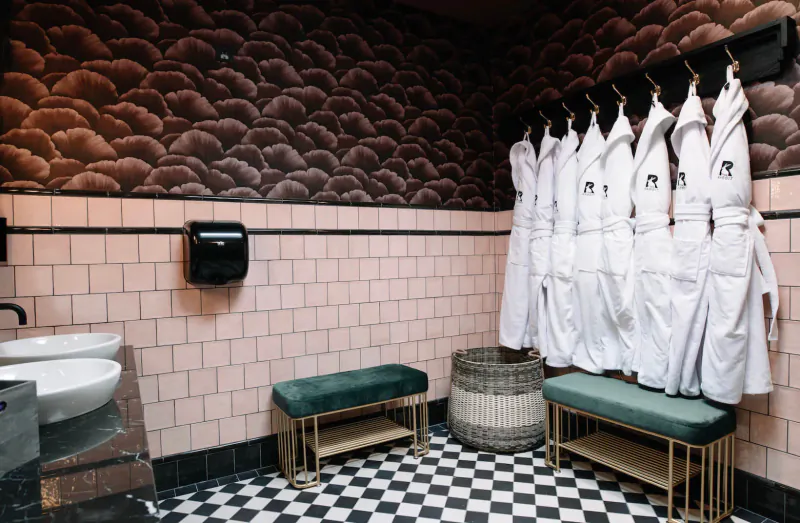 Stylish hotel bathroom with pink tiles, black-and-white checkered floor, white robes on rack, green bench, sinks.