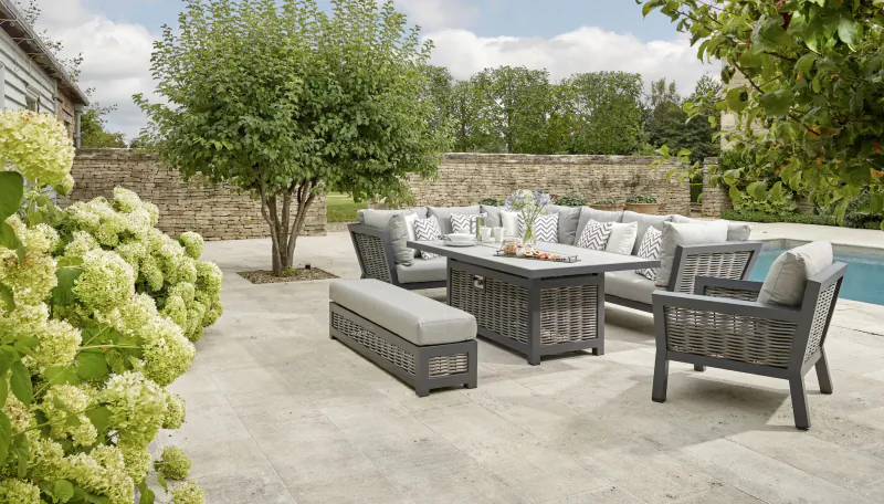 Grey Bramblecrest premium garden lounge set with fire pit table, cushions, bench on tiled patio by pool amid hydrangeas and trees.