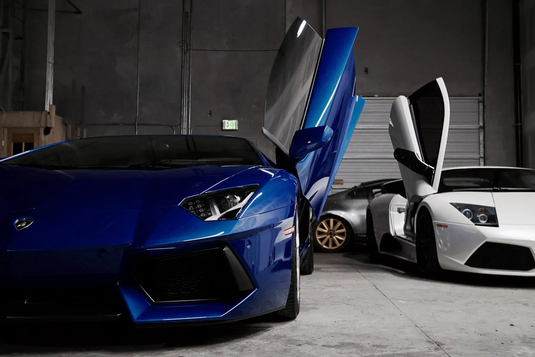 Blue Lamborghini Aventador with doors up beside white Lamborghini in industrial garage