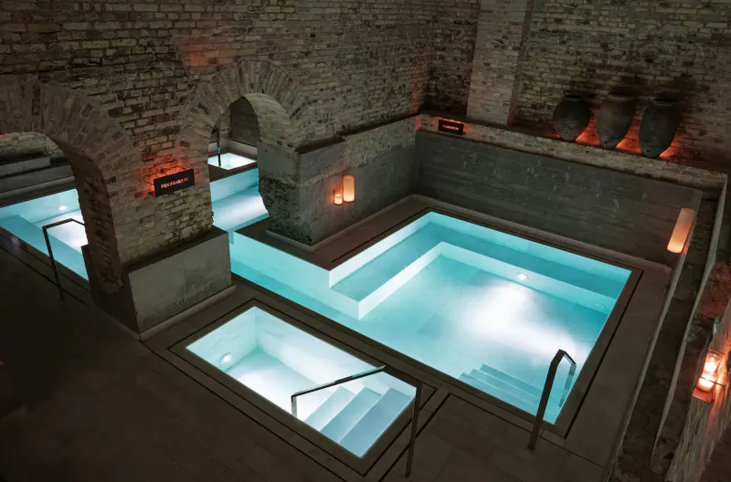 Aerial view of candlelit infinity pools in brick-arched ancient baths, glowing turquoise water and steps.