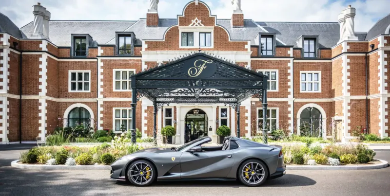 Silver Ferrari sports car parked in front of grand brick chateau entrance with ornate 'F' canopy and gardens.