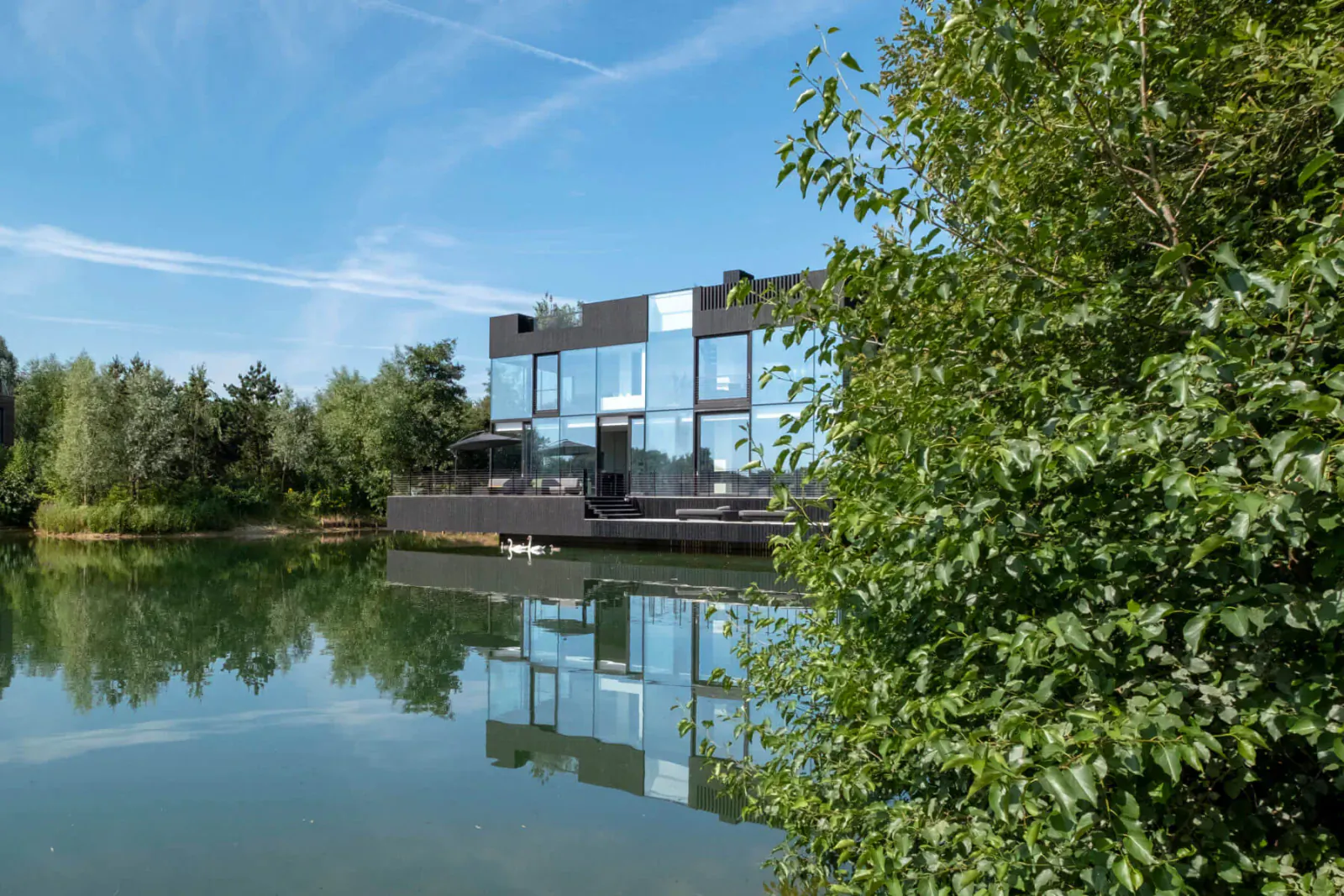 Modern black glass house on lake edge, reflected in calm water amid green trees under blue sky