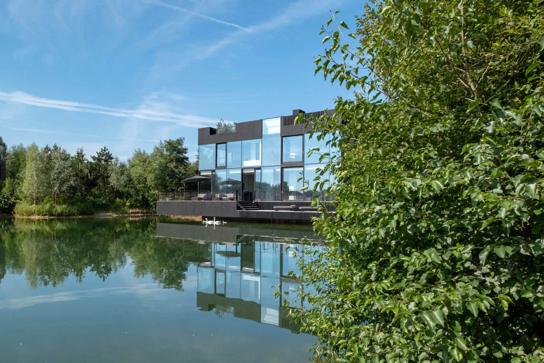 Modern black glass house on lake edge, reflected in calm water amid green trees under blue sky