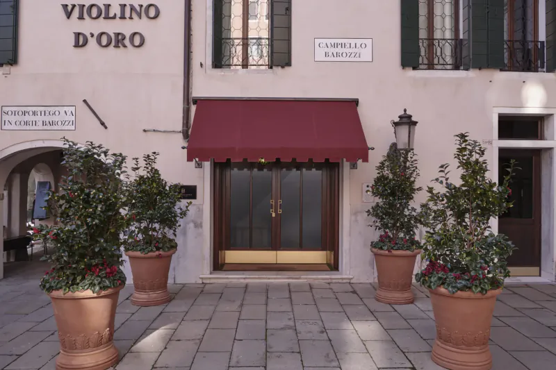 Violino d'Oro Hotel entrance in Venice, with red awning, potted plants, and 'Campiello del Piovan' sign on beige building.