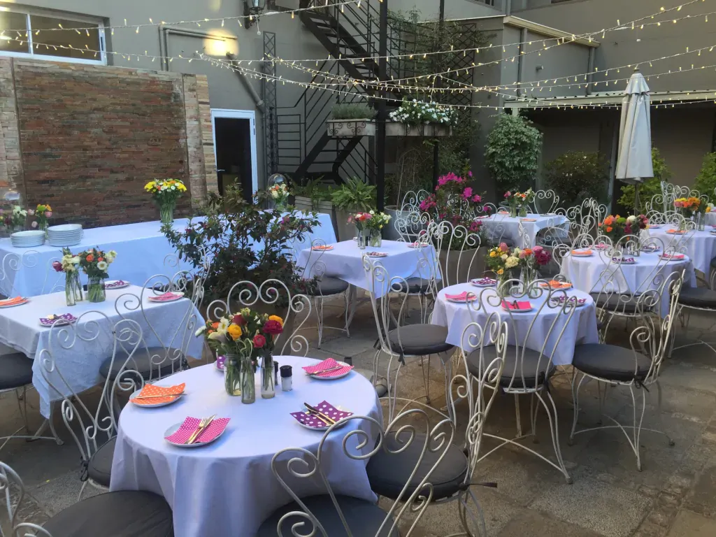 Elegant outdoor courtyard at Arbour Café & Courtyard with white-clothed tables, colorful flower arrangements, string lights, and ornate chairs.