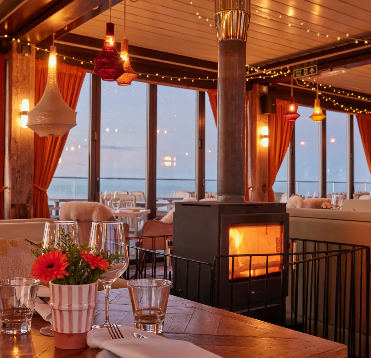 Cozy seaside restaurant interior at Rockwater Hove with lit fireplace, sunset ocean view, pendant lights, orange curtains, and table set with wine glasses and gerbera daisies.