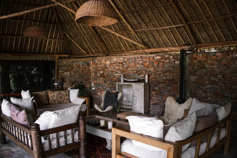 Cozy thatched-roof lounge in Kenyan wild house with white cushions on wooden chairs, woven lamps, brick walls.