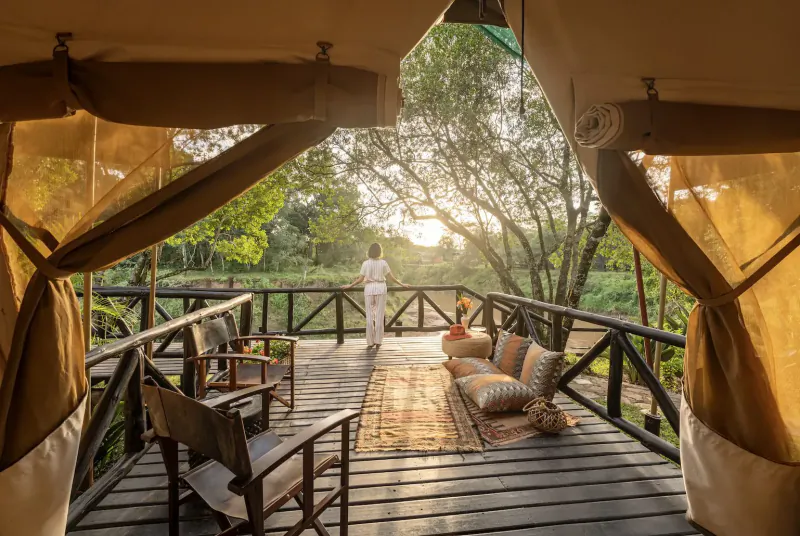 Person in white stands on wooden deck of luxury tent overlooking lush green trees at sunset, with chairs and cushions.