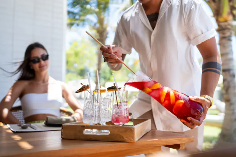 Man in white shirt pours orange sangria from red pitcher into glasses with fruit garnishes on wooden tray at tropical resort table