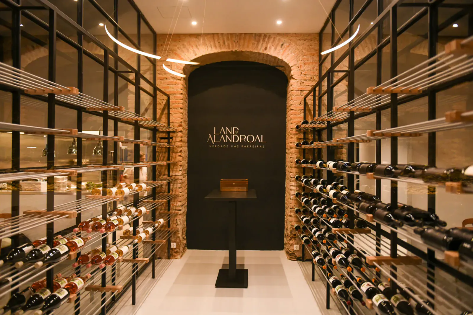 Elegant wine cellar with arched brick wall displaying 'Land of Alandroal' logo, metal racks of wine bottles, and central black pedestal table.