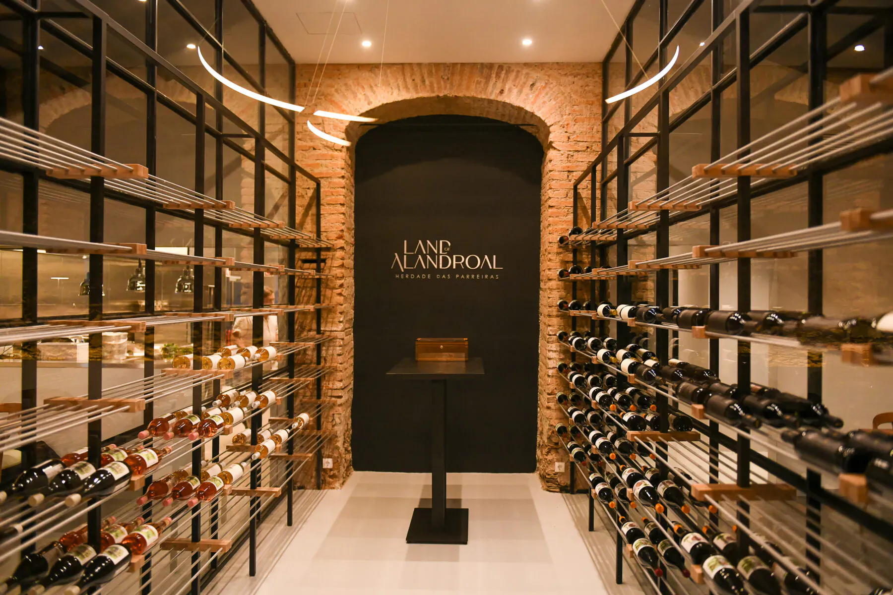Elegant wine cellar with arched brick wall displaying 'Land of Alandroal' logo, metal racks of wine bottles, and central black pedestal table.