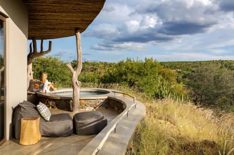 Outdoor hot tub and lounge seating on wooden deck at Mashatu Euphorbia eco camp, overlooking African savanna under cloudy sky.