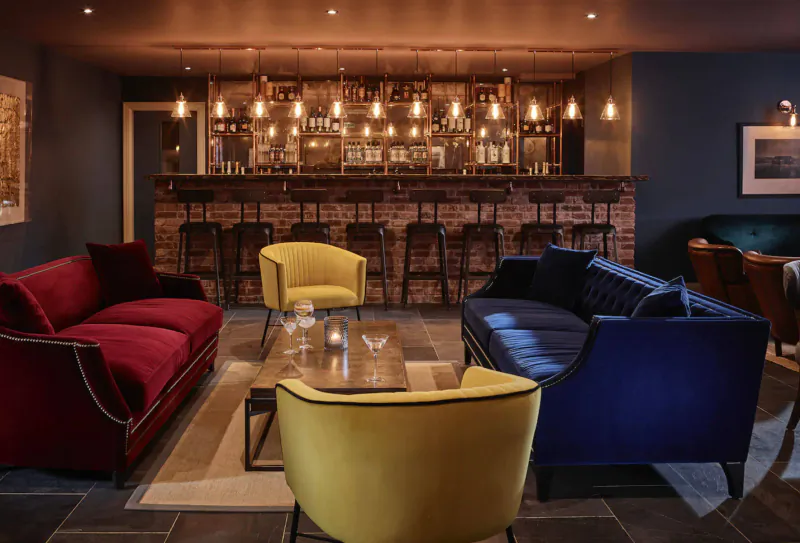 Cozy lounge at The Harper Norfolk with brick bar, pendant lights, yellow and red velvet seating, and coffee table.