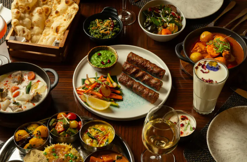 Assortment of Indian dishes on wooden table: naan, kebabs, curries, dal, rice, veggies, lassi, and wine glass.