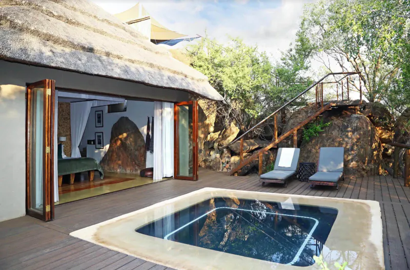 Madikwe Hills lodge suite with thatched roof, open doors to bedroom, hot tub, loungers on wooden deck amid rocks and trees