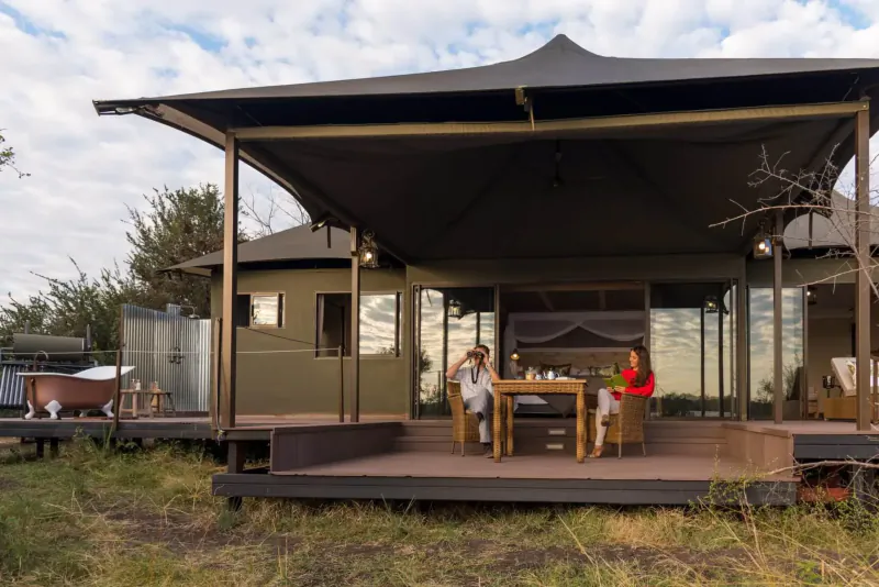 Couple in white and red attire seated at table on deck of luxury safari tent lodge by riverside, with bathtub nearby.