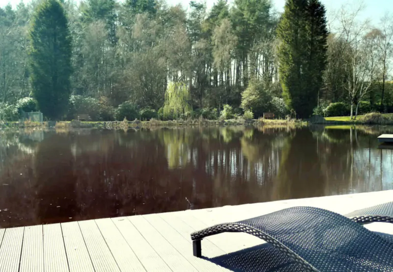 Reclining chair on wooden deck by tranquil pond reflecting tall trees at Moddershall Oaks Spa.