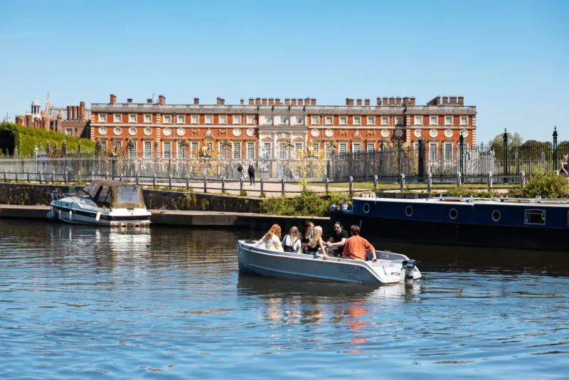 Group in silver motorboat on river with docked boats, red brick palace and gardens in background, clear blue sky