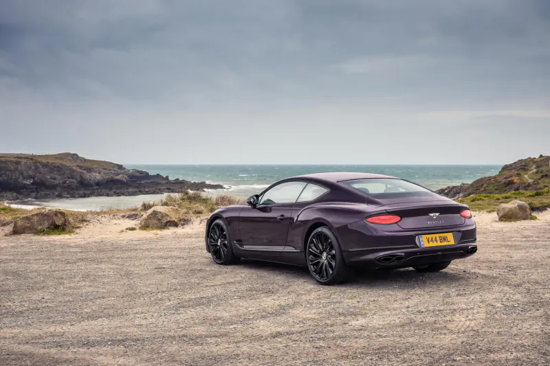 Rear view of purple Bentley GT Mulliner Blackline on gravel beach by ocean, cloudy sky, UK plate.